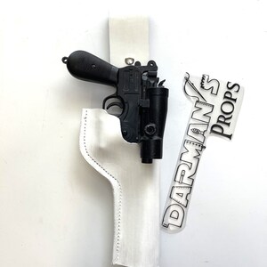 May include: A black plastic toy blaster with a red laser sight in a white leather holster. The holster has a white stitching detail. A sticker with the text "DARMAN'S PROPS" is on the white background.