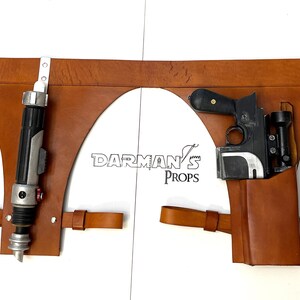 May include: A brown leather belt with two holsters. The left holster holds a black and silver lightsaber. The right holster holds a black and silver blaster pistol. The belt has a metal buckle and a metal loop on the right side. The text "DARMAN'S PROPS" is printed on the belt.