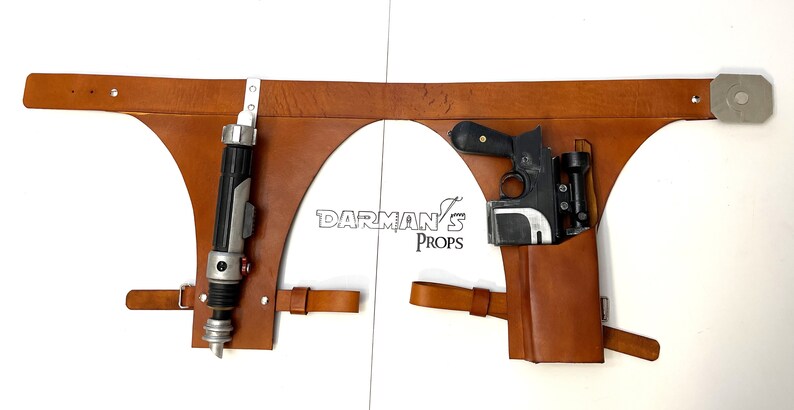 May include: A brown leather belt with two holsters. The left holster holds a black and silver lightsaber. The right holster holds a black and silver blaster pistol. The belt has a metal buckle and a metal loop on the right side. The text "DARMAN'S PROPS" is printed on the belt.