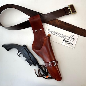 May include: A brown leather holster with a black toy gun and a brown leather belt with a brass buckle. The holster has a strap and buckle. The belt is long enough to fit around a waist. The text "Darman's Props" is visible on a white background.