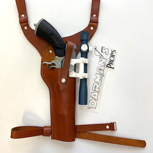 Op de afbeelding: Een bruine leren holster met een schouderriem voor een speelgoedpistool. De holster heeft een metalen gesp en een metalen drukknoop. Het pistool is zwart en zilver met een blauwe scope. De holster is gelabeld "Darman's Props".