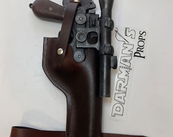 Op de afbeelding: Een rekwisieten blasterpistool met een vizier, genesteld in een bruin leren holster. De holster heeft een riemlus en een riem met druksluitingen. Het pistool is donkergrijs met een bruine grip. Een sticker met de tekst "DARMAN'S PROPS" is zichtbaar.