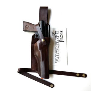 May include: A brown leather holster with a strap for a gun. The holster is designed to be worn on the hip. The holster is empty, but there is a gun in the background.