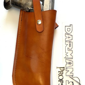 May include: A brown leather holster with a silver gun inside. The holster has a strap with a snap closure. The holster is labeled "DARMAN'S DROPS".