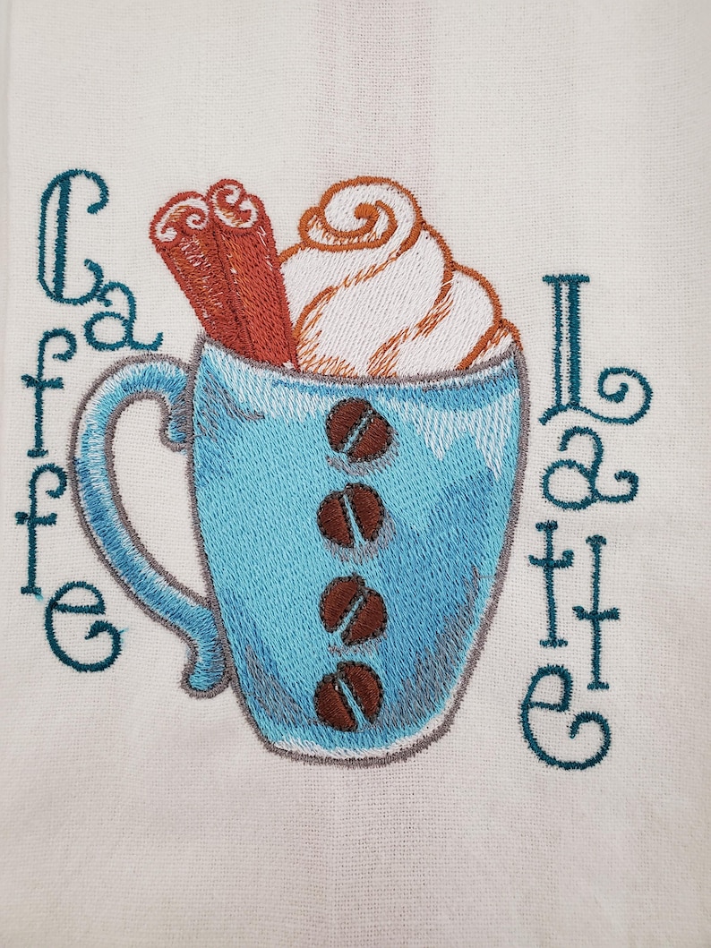 May include: Embroidered design of a blue coffee cup filled with whipped cream, cinnamon sticks, and coffee beans. The words "Cafe Latte" are embroidered in teal on a white background. A cozy, coffee-themed design.