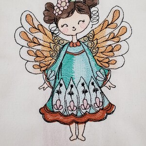 May include: Embroidered image of a smiling fairy with brown hair, a flower crown, and large, detailed wings. The fairy wears a teal dress with floral accents and a brown trim. The artwork is on a white fabric background.