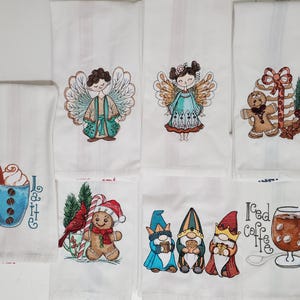May include: White kitchen towels with embroidered designs. The designs include a blue coffee cup with latte art, angels, gingerbread men, gnomes, and iced coffee. The towels are likely for the holiday season.