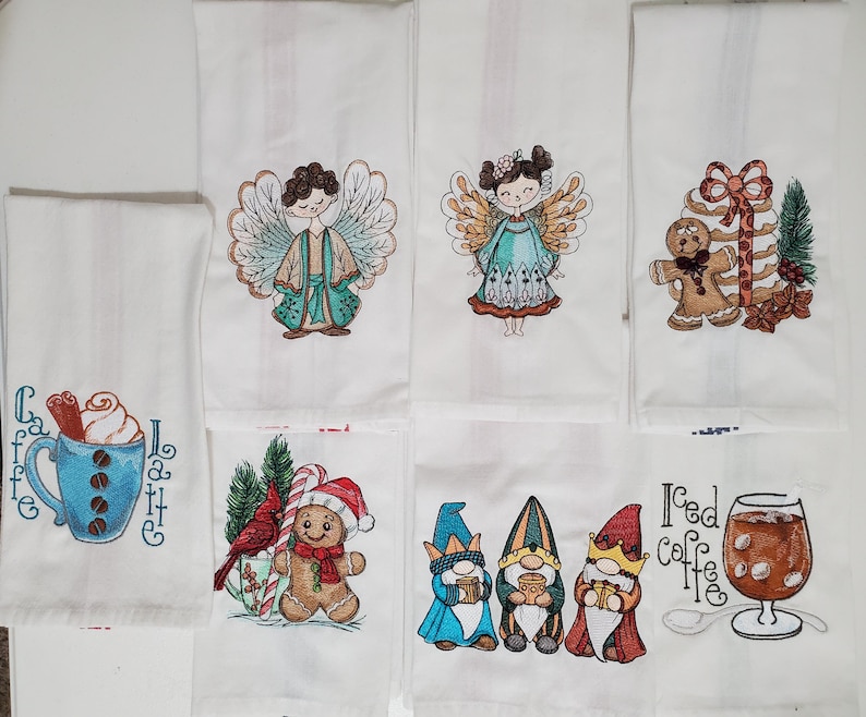 May include: White kitchen towels with embroidered designs. The designs include a blue coffee cup with latte art, angels, gingerbread men, gnomes, and iced coffee. The towels are likely for the holiday season.