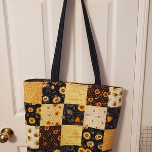 May include: A patchwork tote bag with black straps, featuring a design of sunflowers, bees, and honeycomb patterns in yellow, black, and white. The bag is hanging on a white door.