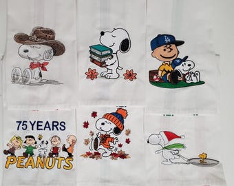 May include: White cotton kitchen towels with embroidered Snoopy designs. Snoopy is depicted in various scenes, including wearing a cowboy hat, holding books, and with Charlie Brown. Other towels feature the Peanuts gang and the text "75 YEARS PEANUTS."