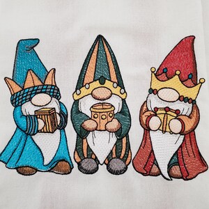 May include: Embroidered image of three gnomes, each wearing a different colored hat and holding a gift. The gnomes have long white beards and are dressed in blue, green, and red robes. The image is on a white background.