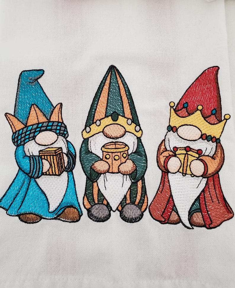 May include: Embroidered image of three gnomes, each wearing a different colored hat and holding a gift. The gnomes have long white beards and are dressed in blue, green, and red robes. The image is on a white background.