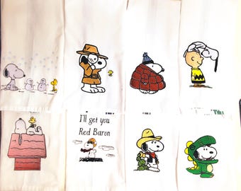 May include: Set of eight white kitchen towels with embroidered images of Snoopy from Peanuts. The towels feature Snoopy in various poses and settings, including a winter scene, a detective, a cowboy, and a dinosaur. The towels are all white with black embroidery.