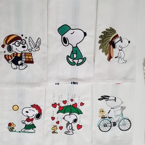 May include: White cotton kitchen towels with embroidered Snoopy designs. Each towel features a different Snoopy character in various outfits and scenes, including a scarf, hat, and a bicycle. The towels are a charming addition to any kitchen.