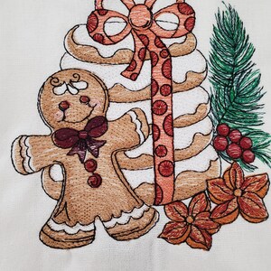 May include: Embroidered Christmas design featuring a gingerbread man with a bow tie, a stack of cookies, a bow, and a sprig of evergreen. The design also includes two orange flowers and red berries. The embroidery is on a white background.