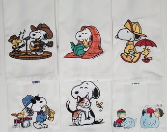May include: White cotton towels with embroidered Snoopy and Woodstock designs. One features Snoopy playing guitar with Woodstock singing, another has Snoopy reading under a blanket, and others show Snoopy with a cat, playing the saxophone, and in the rain.