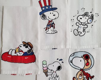 May include: White cotton kitchen towels with embroidered Snoopy designs. Snoopy is depicted in various scenes, including wearing an Uncle Sam hat, listening to music, relaxing in a float, eating ice cream, and wearing a space helmet. Each towel has a striped border.
