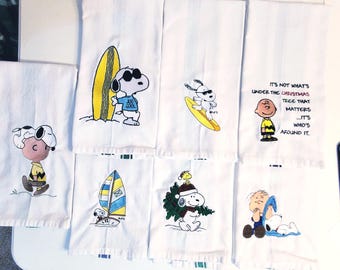 May include: Set of six white kitchen towels featuring Peanuts characters. The towels have different designs, including Snoopy surfing, Charlie Brown with a Christmas tree, and Snoopy with a sailboat. The towels have black embroidered text that reads "It's not what's under the Christmas tree that matters - It's who's around it."