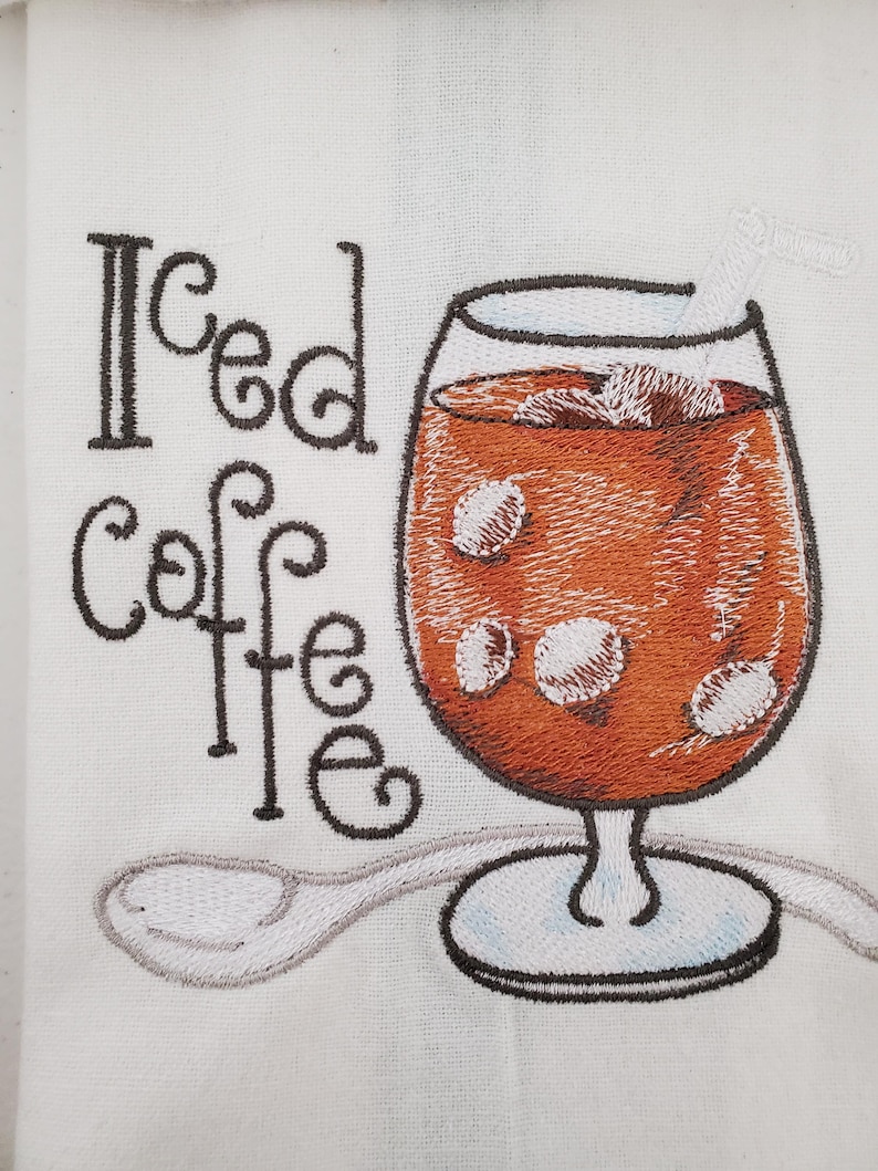 May include: Embroidered white fabric features a glass of iced coffee with ice cubes and a straw. The words "Iced Coffee" are embroidered in black script. A spoon is embroidered below the glass.