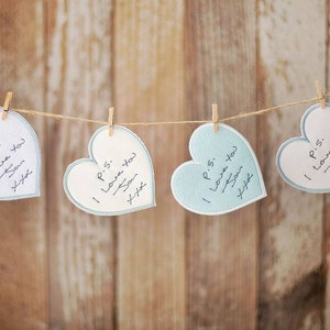 May include: Four heart-shaped cards with handwritten messages, suspended from a string with pegs. The cards alternate between white and light blue, with the text "P.S. I Love You Son xxx". The background is a wooden surface.