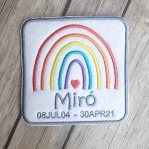 May include: A white square patch with a rainbow design and a red heart in the centre. The text "Miró 08JUL04 - 30APR21" is embroidered in grey below the rainbow.