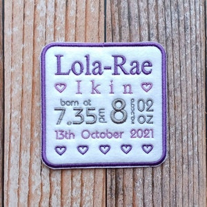 May include: A white square patch with purple stitching, embroidered with the text "Lola-Rae" in purple, "I kin" in pink, "born at 7.35" in grey, "8 lb 02 oz" in grey, and "13th October 2021" in purple. There are small pink hearts embroidered around the text.
