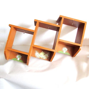 May include: A wooden wall shelf with three compartments. Each compartment has a painted floral design. The shelf is brown and has a rustic look.