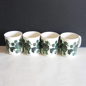 May include: Four white ceramic mugs with a dark green floral pattern. Each mug has a cylindrical shape with a handle on one. The floral design includes stylized flowers and leaves. The mugs are arranged on a white surface against a dark background.
