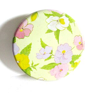 May include: A round, green tin with a floral pattern. The tin is decorated with pink, white, and purple flowers with green leaves.