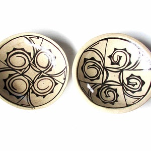 May include: Two small ceramic bowls with a white background and black swirling designs. The bowls are round and have a slightly raised rim.