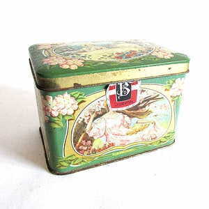 May include: A vintage green metal tin with a floral design and a woman in a pink dress. The tin has a label that says "B of DEVON" and "PRIMO".