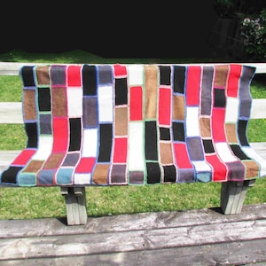 May include: A colorful patchwork quilt draped over a wooden bench. The quilt features rectangular blocks in red, black, white, brown, and blue. The bench is made of weathered wood and sits in front of a white fence and green grass.