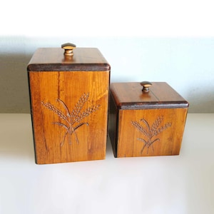 May include: Two wooden storage boxes with wheat stalk carvings. The boxes are rectangular with dark brown lids and knobs. The wood has a warm, light brown tone. The larger box is taller than the smaller box. The wheat stalk design is carved into the front of each box.