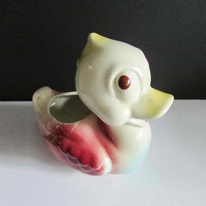 May include: A ceramic duck planter with a white body, yellow beak, and red eye. The duck is painted with pink and blue accents.