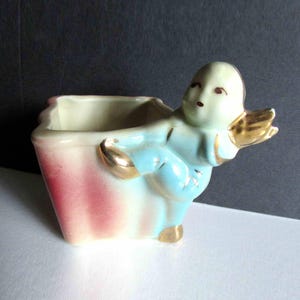 May include: A vintage ceramic planter in the shape of a cherub. The planter has a square base with a pink and white striped design. The cherub is light blue with gold accents on the wings and feet. The interior is cream-colored.