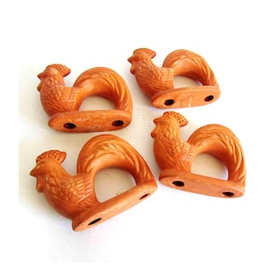 May include: Four terracotta-colored rooster-shaped napkin rings. Each ring features a detailed rooster design with a curved body and a base with two holes. The napkin rings are arranged on a white background.