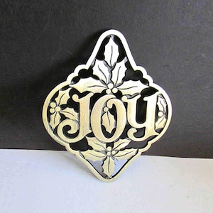 May include: A gold-colored metal ornament with the word "JOY" in a decorative font. The ornament is shaped like a diamond with holly leaf and berry designs around the edges. The background is black and white.