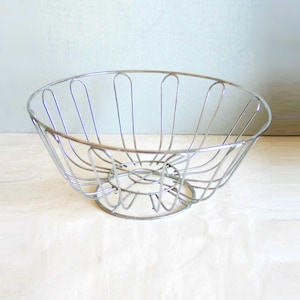 May include: A silver-colored wire fruit basket. The basket has a rounded shape with vertical bars and a circular base. The design is open, allowing for air circulation. The basket is empty and sits on a light-colored surface.