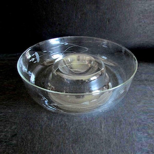 May include: A clear glass bowl with a smaller, circular bowl in the center. The glass has etched floral designs. The bowl is likely used for serving dips or snacks. The bowl is on a dark surface.