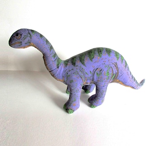 May include: A plush dinosaur toy in shades of purple and green. The long-necked dinosaur has a detailed design with green spots and accents. The toy is standing on a white surface.