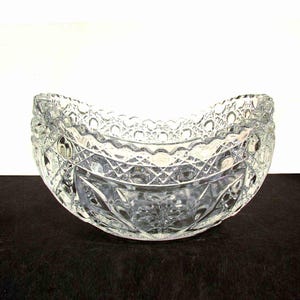 May include: A clear, decorative glass bowl with a scalloped rim and intricate diamond and floral patterns. The bowl is crescent-shaped and sits on a dark surface, showcasing its detailed design.