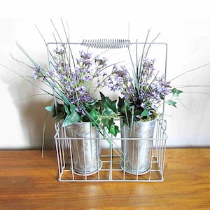 May include: Two silver metal flower pots with purple flowers and green leaves in a silver wire basket with a handle.