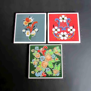 May include: Three square tiles with painted floral designs. The first tile has a blue background with a red, white, and yellow floral design. The second tile has a red background with a white, blue, and yellow floral design. The third tile has a green background with a yellow, white, and orange floral design.