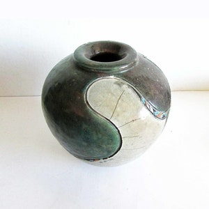 May include: A round ceramic vase with a green and white glaze. The vase has a unique design with a curved shape and a small opening at the top.