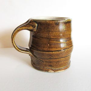 May include: A handmade brown ceramic mug with a large handle. The mug has a textured, earthy appearance with horizontal striations and a lighter interior. The mug is approximately 4 inches tall.