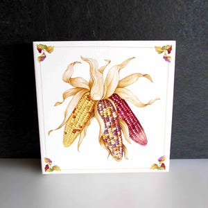 May include: Square ceramic tile featuring a watercolor illustration of three ears of corn. One is yellow, one is purple and white, and one is red. The husks are tan, and the corners have floral accents.