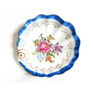 May include: A white porcelain dish with a blue and gold rim. The dish is decorated with a floral design in shades of pink, purple, and blue. The dish has a scalloped edge and a small handle.