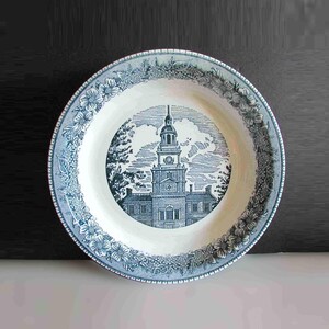 May include: A round, white and blue decorative plate. The plate features a detailed illustration of a building with a clock tower in the center. The rim of the plate is decorated with floral and foliage patterns. The plate is a vintage style.