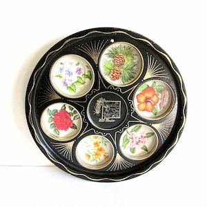 May include: A black metal tray with a gold rim and six circular compartments. Each compartment features a different floral design, including a rose, a hibiscus, and a pine cone. The center compartment features a map of New Mexico with the text "New Mexico".