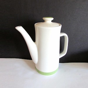 May include: A white ceramic coffee pot with a long spout, handle, and lid. The pot has a green band around the base and a green knob on the lid. The background is black and white.